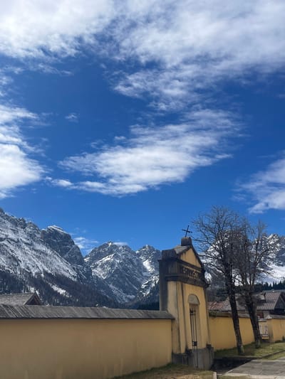 The image shows a view of a church bell tower in Sappada/Plodn/Sapade, Italy, set against a backdrop of snow-capped mountains and a blue sky with scattered clouds. The tower is a light yellow color, with a cross at its peak and the word "RESURRECTURUS" inscribed on its side. A bare tree stands nearby. Part of the adjacent building is visible, with some stone and wooden trim. The mountains rise in the background, partially covered with snow, and the sky is clear with wispy white clouds.