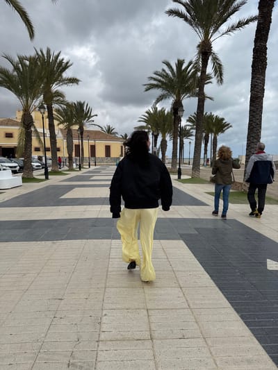 The image displays an outdoor scene on a tiled promenade in Los Alcázares, Spain, under an overcast sky. The foreground features a paved pathway composed of alternating light and dark grey rectangular tiles. A person, viewed from behind, walks away from the camera in the center. This individual wears a black bomber jacket, loose-fitting light yellow wide-leg pants, and dark shoes; their long, dark, wavy hair is visible. To the right, two additional individuals are present: a person with curly brown hair, wearing a dark olive green jacket and blue jeans, is seen from behind looking towards the right, and another person in a grey and dark blue jacket with dark pants walks ahead. Numerous tall palm trees with green fronds and dark trunks line both sides of the promenade, interspersed with dark streetlights. In the background on the left, several light yellow buildings with dark roofs and shutters are visible, with multiple white parked cars in front of them. To the right in the midground, a sandy beach leads to a large body of water extending to the horizon under the grey clouds.