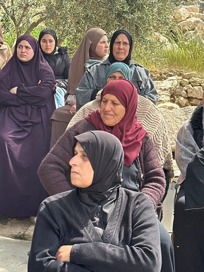 The image depicts a group of at least seven women gathered outdoors in Dura, Palestinian Territory. The women are dressed in traditional Islamic attire, including various colored hijabs (head coverings) and modest clothing. In the immediate foreground, a woman with a black hijab and ribbed black cardigan is seen in profile, looking left with her arms crossed. Directly behind her, an older woman with a dark red hijab and textured maroon top looks downwards and slightly right. To the far left, a woman in a dark purple abaya and hijab stands with crossed arms, looking towards the left foreground. Behind her, a younger woman in a black jacket and black hijab looks forward. To her right, a woman in a brown hijab and a grey patterned jacket looks right, while an older woman next to her, wearing a black hijab and a similar grey patterned jacket, looks forward. A partial view of a woman with a teal hijab is visible behind these two. The background features rocky terrain with sparse green vegetation and what appears to be olive trees under daylight conditions. The women appear to be observing something off-camera, with varied expressions and postures.