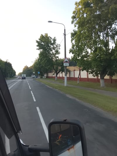 The image depicts a view from inside a vehicle driving on a road in Zhlobin, Belarus. The road has multiple lanes and is marked with white lines. On the right side of the road, there is a sidewalk, a grassy area, and a brick wall with a white stucco finish. Trees line the roadside, with one large tree prominent in the foreground. A street light is visible on the right side of the road, and a traffic sign indicating a speed limit of 40 km/h and a road hazard (speed bump) is mounted on a pole next to it. A pedestrian crossing is marked on the road ahead. Other vehicles, including cars and a truck, are visible on the road in the distance. The sky is clear, suggesting daytime, possibly late afternoon or early evening given the soft light. The overall scene appears to be a suburban or semi-rural area. There is no explicit indication of people engaged in any specific activity, nor any discernible emotions. The time of day appears to be late afternoon or early evening due to the light.