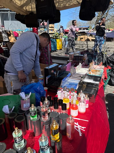 The image captures a scene at an outdoor market. A vendor, dressed in a gray hoodie and jeans, sits behind a table draped with red fabric, which displays an assortment of perfumes and fragrances in various bottles and packaging. To the vendor's left, a row of black bags hangs from a rack. In the background, people stroll along a walkway beside a flight of stairs, suggesting a moderately bustling environment. The products on the table feature labels like "LAXURIA," "OUD VELVET," "FRAISE," and "COLLECTION PRI." Based on the visible clues, the market appears to be located in Garges-lès-Gonesse, France.