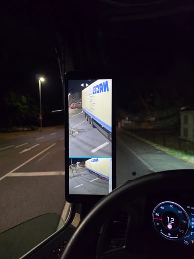 This is a nighttime, interior shot from the cabin of a truck, looking forward. A large, dark steering wheel dominates the foreground, with a digital dashboard displaying a speed of 12 km/h. To the left of the steering wheel, a vertical tablet-like screen is mounted, showing two camera feeds. The top feed displays a yellow truck with blue lettering that appears to read "HERM." The truck is parked in what looks like a parking lot or service area. The bottom feed shows a similar scene with the same truck. Outside the cabin, through the windshield, a dimly lit road with white lane markings is visible, along with a streetlamp and trees. The environment suggests an urban or semi-urban setting in Flörsheim, Germany, likely at a truck stop or logistics area. There are no people visible in the image.