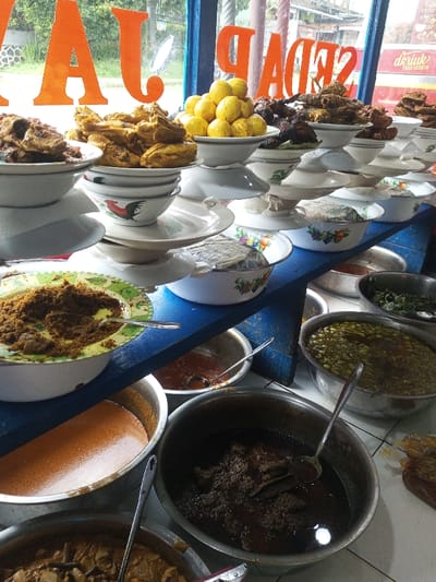 A variety of Indonesian dishes are displayed in bowls and on stacked plates. Dishes include beef rendang, curried chicken, boiled eggs, and various vegetable dishes. The food is arranged on a blue countertop, with visible signs that spell out "JAJA SEDAP" in orange. Spoons are placed in several of the dishes. A window is behind the countertop.