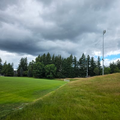This media file depicts a grassy field, likely a sports field, in Portland, United States. The scene is dominated by vibrant green grass, neatly mowed in the foreground and transitioning to taller, more wild grass further up a gentle slope. The field appears to be part of an outdoor recreational area, possibly a park or school grounds, given the presence of a soccer goal on the left side of the frame, partially obscured by trees.

Behind the field, a dense line of coniferous trees forms a natural backdrop, with some deciduous trees interspersed. The sky above is a mix of dramatic, dark storm clouds and patches of bright blue sky with fluffy white clouds, suggesting an unsettled weather pattern. The presence of a tall light pole with multiple fixtures on the left side of the frame indicates the field is equipped for nighttime use. Another, shorter light pole is visible further back, also near the trees.

There are no people visible in the image, and no specific activity is taking place. The image seems to capture a moment of natural beauty and the potential for outdoor recreation, juxtaposed with the approaching or receding weather. The overall mood is one of tranquility and the vastness of nature. No visible text is present in the image. The time of day is ambiguous due to the varied light conditions caused by the clouds, but it appears to be daytime.