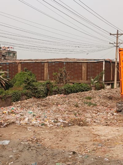 This image depicts a wide-angle view of a heavily littered urban landscape in Boliarpur, Bangladesh, under an overcast sky. The foreground is dominated by an extensive, irregular mound of mixed waste, primarily consisting of plastic bags, bottles, and assorted refuse, covering a dirt ground with scattered debris and dry organic matter. No individuals are present.
In the midground, a low, weathered brick wall runs horizontally, topped by dense, unruly green vegetation, including bushes and vines. Behind this foliage stands a long, single-story building constructed from vertically oriented, heavily rusted corrugated metal sheets. Its flat roof, also appearing to be made of corrugated material, shows signs of weathering.
The background features a multi-story, light-colored residential or commercial building on the left, with laundry visible hanging on its rooftop. Numerous black utility lines crisscross the upper portion of the frame against a uniform light grey sky, originating from a utility pole partially visible on the far right. An orange shipping container is positioned vertically on the far right edge of the frame, adjacent to the utility pole.