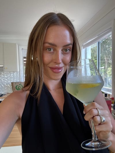 A woman holds a cocktail glass filled with a light-colored drink and a lemon slice. She wears a black top and has rings on her fingers. Her hair is light brown and styled, and she has green eyes. Behind her, a window shows greenery. The background includes a white wall, tiled cabinets, and a wooden surface.