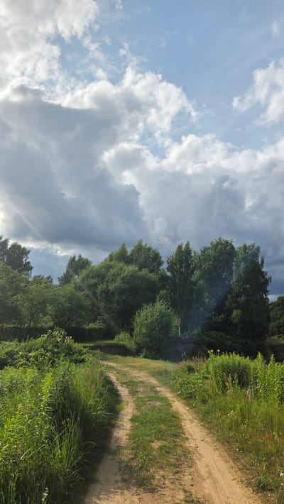 The image depicts a rural path winding through lush green vegetation on the right side, with a dense forest bordering the path further in the distance. The sky on the left is a mix of bright blue and dramatic, greyish-white clouds, suggesting dynamic weather conditions. Sunlight streaks through the clouds, illuminating parts of the scene with a soft, hazy glow. The path itself appears to be made of sand or dirt, with visible tire tracks, indicating it's a frequently used route, likely for vehicles or bikes. The overall scene evokes a sense of natural beauty and tranquility, with the vibrant greenery and expansive sky creating a picturesque landscape. The time of day appears to be late afternoon or early evening, given the angle of the light and the slightly muted tones in some areas, with the weather being partly cloudy and bright. The location is Cekule, Latvia. There is no visible text in the image.