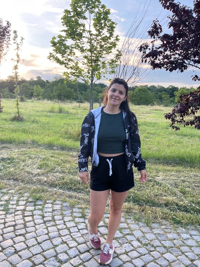 A young woman stands upright on a grey cobblestone path, facing slightly left and smiling gently at the camera. She has dark hair and is dressed in a dark green crop top, black shorts with a white drawstring, and a black and white jacket featuring the word "Angel" printed multiple times on its sleeves. She wears maroon or pink sneakers with white laces. To her left, a wide expanse of green grass leads to a line of trees in the background. The sky above is bright with scattered clouds, displaying the warm, soft light of late afternoon or early evening, possibly near sunset. Tree branches with both dark red and green leaves are visible in the foreground, framing the outdoor scene. The overall setting appears to be a park or natural recreational area.