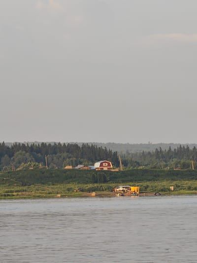 The image shows a wide river in the foreground, reflecting the overcast sky. On the far bank, a verdant slope is covered with grass and vegetation, dotted with trees. A cluster of buildings is situated atop the slope, including a prominent red barn-like structure with white trim and a small window. Further along the bank, several other houses and structures are visible, some with peaked roofs. Behind these buildings, a dense forest of dark green trees stretches across the horizon. On the riverbank in the middle ground, several objects are present, including what appears to be a yellow modular structure, a white vehicle, and a dark-colored car, as well as a small boat. The overall scene suggests a rural or semi-rural setting, possibly a settlement along a river. The location is identified as Lesosibirsk, Russia.