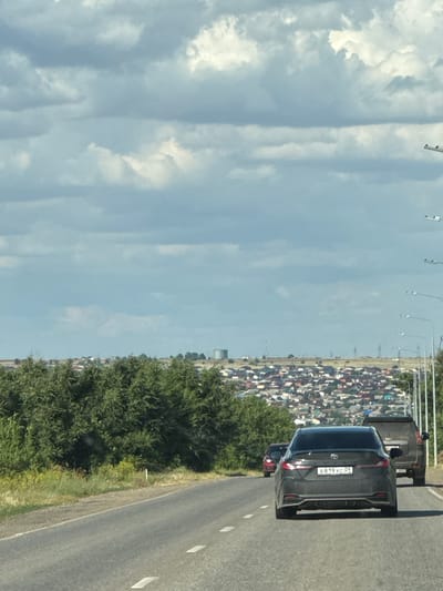 This is a landscape photograph taken from a vehicle moving on a road, oriented vertically. The foreground shows the road surface with white lane markings. To the left of the road is a dense green tree line, and beyond that, a vast expanse of sky filled with scattered white clouds.
In the background, on the right side of the road, a long line of buildings stretches into the distance. The buildings appear to be mostly residential, with a variety of roof colors, creating a dense urban or suburban landscape. Several cars are visible on the road, including a dark-colored SUV in the middle ground, displaying a license plate that reads "B819XC 09". Further ahead, a very long queue of vehicles is visible, suggesting a traffic jam or a procession of some kind. Beyond the buildings, the horizon is hazy.
The scene suggests daytime, with bright sunlight illuminating the clouds and the road. The weather appears to be clear and sunny. The presence of the license plate with Cyrillic characters indicates the location is likely Russia. The overall impression is a snapshot of a journey through a populated area on a sunny day.