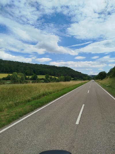 This media file depicts a scenic view of a rural road in Boxberg, Germany. The scene is dominated by a grey asphalt road with white dividing lines, stretching into the distance. The road is bordered by lush green grass and tall, golden-brown grass on either side. In the background, a dense forest covers a rolling hill, with patches of yellow fields visible behind the trees. The sky above is a vibrant blue, adorned with fluffy white clouds, suggesting a pleasant day.
A white car is visible in the distance, traveling on the road. The lighting suggests it is daytime, likely late morning or early afternoon, given the bright sunlight and clear sky. The overall atmosphere is peaceful and serene, evoking a sense of travel and exploration through the German countryside. There are no visible people or text in the media file. The location cues such as the road markings, the types of trees and vegetation, and the rolling hills are consistent with a European landscape, specifically Germany.
