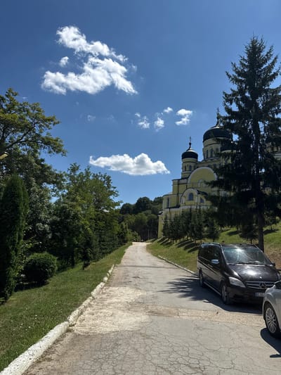 The image depicts the Monastery of Hancu in Ciuciuleni, Moldova, on a sunny day with a clear blue sky dotted with white clouds. A paved road, cracked and worn, leads away from the viewer and towards the monastery. To the left of the road, a grassy slope is covered with trees and bushes, including a tall cypress tree.

Dominating the right side of the frame is a portion of the monastery's yellow facade, characterized by its multiple domes and intricate architectural details. In the foreground, a black minivan is parked on the right side of the road, partially obscuring the view of a silver car parked behind it. Shadows from trees are cast across the road and the vehicles.

The overall impression is a serene, somewhat rustic scene at a significant religious site, with natural elements and human presence in the form of vehicles.