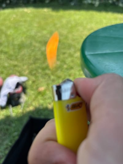 A person's hand is holding a yellow BIC lighter outdoors. The lighter is in focus, while the background is blurred. The background shows a grassy area, a green table, and some indistinct objects. An orange flame is visible emanating from the lighter. The scene appears to be a casual outdoor setting. There is no information in the image to suggest the location is Vicenza, Italy.
