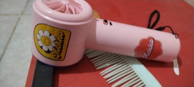 In this medium shot, a pink, novelty hairdryer, designed to resemble a cartoonish face, is laid on a red surface, possibly a table. The hairdryer has a round, fan-like opening at one end, and a cylindrical body with a small, black strap attached to the back. A yellow sticker with a smiling daisy and the word "Mekko" in a bubbly font is adhered to the side of the hairdryer. Further down the body of the hairdryer, a red cloud-shaped sticker also displays the "Mekko" logo.
Beneath the hairdryer, a light blue comb with thin teeth is partially visible, its bristles fanned out. The red surface contrasts with the lighter tones of the hairdryer and comb, and a tiled floor can be seen in the background on the left. The lighting suggests an indoor setting, and the overall impression is of a collection of personal care items. There are no people or animals in the image, and no specific activity is clearly depicted, other than the items being placed on the surface. The location context of Duman, Indonesia, provides a geographical anchor without adding visual cues to the image itself. The visible text consists of the brand name "Mekko" appearing on two stickers.