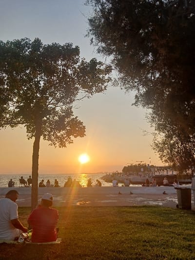 The image depicts a waterfront scene in Karşıyaka, Turkey, at sunset. In the immediate foreground, two individuals are seated on a grassy patch, seen from behind. The person on the left wears a white shirt and light trousers, while the person on the right wears a red shirt and a white baseball cap. Objects resembling a foil-wrapped item and a green container are on the grass next to the individual in white. To the left, a small, silhouetted tree stands.
The mid-ground features a wide concrete promenade or path, appearing wet and reflective in areas, separating the grassy area from the water. Numerous individuals, approximately fifteen to twenty, are dispersed along the concrete edge, most facing the setting sun. Some are seated on portable chairs, others directly on the concrete barrier. To the far right of the promenade, a dark, rectangular refuse bin is visible.
In the background, a large body of water, consistent with Izmir Bay, extends to the horizon. The sun, a bright orange orb, is positioned low on the horizon, creating a prominent sun flare that reflects across the water. The sky transitions from vibrant orange and yellow hues near the horizon to a lighter blue overhead. Distant landmasses or structures are faintly discernible on the far right horizon. Several small boats or a pier structure are visible further along the coastline to the right. A large, dark-foliaged tree partially frames the scene from the upper right, while the smaller tree on the left also contributes to the framing.