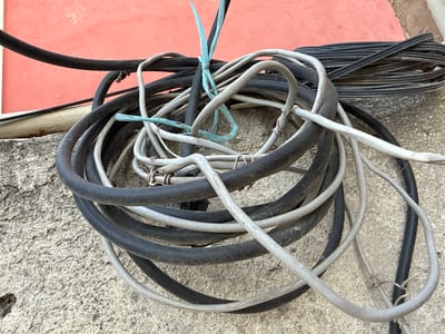 A close-up shot captures a tangled mess of electrical wires coiled on a rough, textured concrete surface. The wires are of various thicknesses and colors, predominantly black and grey, with a bright blue zip tie binding a section of the bundle together. Some wires are thicker and appear to be power cables, while others are thinner and might be data or coaxial cables. They are arranged in a somewhat haphazard heap, with loops and curves creating a visually complex pattern.
In the background, a portion of a textured, reddish-pink wall is visible, suggesting an outdoor setting. The lighting indicates it is daytime, possibly sunny, given the shadows and highlights on the wires and the texture of the concrete. There is no discernible activity or event taking place; the image focuses solely on the static arrangement of the cables.
Notable details include the rough texture of the concrete, which shows some wear and imperfections, and the faded appearance of the colored wall. The wires themselves appear slightly dusty or weathered, indicating they have been exposed to the elements. There is no visible text on the wires or in the immediate surroundings that can be read. The overall impression is one of disarray and utility, a common sight in areas where electrical infrastructure is present. The setting, with the concrete and the painted wall, could be an exterior wall of a building or a similar structure.
