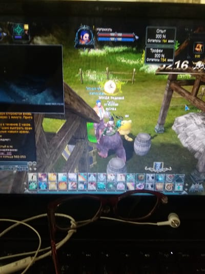 The image shows a screen displaying a video game. On the screen, there is a character in a virtual environment with grass and wooden structures. Various game interfaces with text and icons are visible, indicating player stats and abilities. A pair of red-rimmed glasses and white earbuds are placed in the foreground, obscuring part of the bottom of the screen. The environment depicted in the game does not resemble any specific location in Eshanguzar, Uzbekistan.