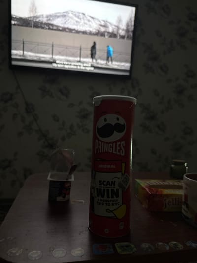 The image displays an indoor domestic setting located in Ciorescu, Moldova, under dim ambient lighting. A flat-screen television is mounted on a wall featuring floral-patterned wallpaper. The TV screen shows a blurred winter scene with snow-covered mountains in the background, a fence in the midground, and two figures walking: one in dark attire on the left and another in a bright blue coat on the right. Subtitles are visible at the bottom of the screen, stating, "Won't Mark, spreading about my open opposition to her regime."
In the foreground, a dark, worn rectangular table with rounded corners is present. Centered on the table is an upright red Pringles 'Original' flavor can, which features the Pringles mascot and a 'SCAN TO WIN A MONOPOLY TRIP TO NYC' promotional label. To the left of the Pringles can, a small, open plastic container, consistent with yogurt, contains a spoon and exhibits a dark label with visible red berries. To the right of the Pringles can, a partial view of a rectangular box labeled 'Jelli' marshmallows is visible, along with a segment of a multicolored ceramic mug. The table surface also displays several circular translucent marks and a line of small square stickers. No individuals are present within the immediate physical scene.
