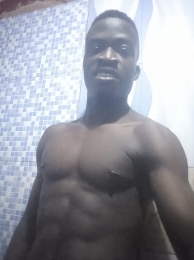 A shirtless, dark-skinned man stands in a brightly lit room. The background features a patterned wall with square tiles on one side and a vertical, wavy design on the other. The man has a muscular physique with visible abdominal muscles and chest definition. He appears to be directly facing the camera. His expression is a slight grimace or forced smile. The lighting is somewhat harsh, creating strong shadows and highlights on his body.