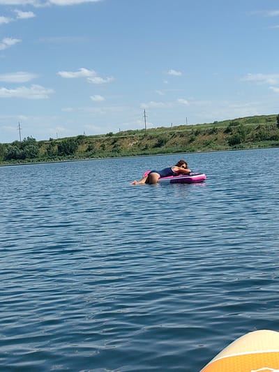 A person is lying on a pink inflatable paddleboard in the middle of a calm body of water. The person is wearing a navy blue swimsuit with white trim and appears to be resting their head on their arms. The paddleboard has some text on it that is not clearly legible. The water is blue with small ripples. In the background, there is a green, hilly shoreline with some trees and what appear to be power lines. The sky is blue with scattered white clouds. The overall scene suggests a warm, sunny day. There is no identifiable landmark to pinpoint the location within the city of Samoilov, Russia, but it is a natural outdoor setting.
