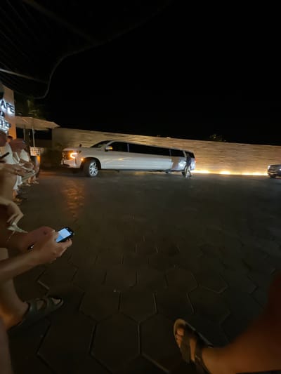 The image depicts a nighttime scene outdoors, with a focus on a group of individuals and a white car.
Environment:
The location appears to be a well-lit urban area, with a building on one side. The ground is paved with dark, hexagonal tiles.
People:
Multiple individuals are present, but not all are clearly visible. One person is holding a mobile phone, seemingly taking a picture. Another is wearing sandals.
Subjects and Objects:
A white car is parked beside the building. A mobile phone is being held up.
Actions:
The person with the mobile phone is likely taking a picture. The rest of the people are socializing or resting in the outdoor area.
The scene resembles a typical social gathering in an urban setting.