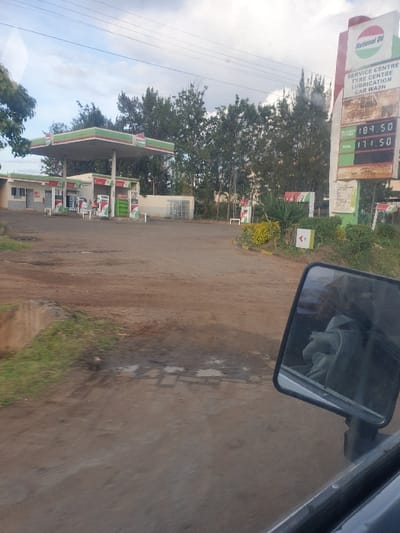 An upright, eye-level shot captures a roadside gas station on a partially unpaved, brownish-grey road, likely in Mumbuni North ward, Kenya. The scene is viewed from inside a vehicle, with the passenger-side mirror visible in the upper right, reflecting a glimpse of the vehicle's interior and a person.
On the left side of the road stands a tall, prominent sign for "NATIONAL OIL," featuring its distinct green and red leaf-like logo. Below the brand name, the sign advertises a "SERVICE CENTRE," "TYRE CENTRE," "LUBRICATION," and "CAR WASH." Below these services, a digital price board displays fuel prices: PETROL at 184.50 and DIESEL at 171.50, with a third, lower price of 11.50, likely for Kerosene.
Further down the road on the left, the green and red canopy of the National Oil gas station is visible, with several fuel pumps beneath it. A white building associated with the station is also seen. The area around the gas station is populated with numerous green trees and bushes, indicating a semi-rural or peri-urban setting. Utility lines run across the upper left portion of the sky.
The sky is largely overcast, suggesting a cloudy daytime, possibly late morning or afternoon. The road surface shows patches of dampness or small puddles, implying recent rain or generally moist conditions. The overall impression is a functional stop along a local route.