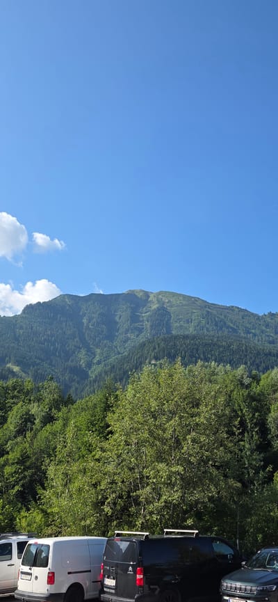 In Sankt Veit im Pongau, Austria, a wide shot captures a mountainous landscape under a clear blue sky with scattered white clouds. The foreground is occupied by a row of parked vehicles, including a white van with a license plate reading "JO 894 GJ" and a black van with a license plate reading "JO 808 BK". Behind the vehicles, a dense forest of green trees dominates the mid-ground. The background features a large mountain, partially covered in trees and with visible ski tracks or paths on its slopes. The overall scene depicts a bright, sunny day in a mountainous region, likely during warmer months given the lush greenery.