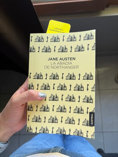 A hand holds a paperback copy of "La Abadia de Northanger" by Jane Austen, published by Austral. The book has a pale yellow cover with a repeating pattern of open books and keys. A yellow bookmark with Spanish text protrudes from the side. The hand has silver glitter nail polish. The background shows a glimpse of jeans and a watch, suggesting the person is seated.