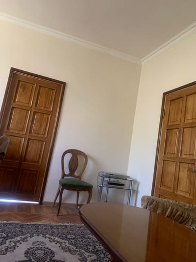 The image depicts an indoor scene, possibly a living room, situated in Sevan, Armenia. The room features two wooden doors, one on the left and another on the right side of the frame, both with a paneled design. In the center of the image, a vintage-style wooden chair with a green upholstered seat stands. To the right of the chair, a small glass and metal shelf unit is visible, holding what appears to be electronic devices. A portion of a polished wooden table is in the foreground, and a patterned rug covers the floor. The walls are painted a light, neutral color, and ornate crown molding is present along the ceiling. The lighting suggests it is daytime.