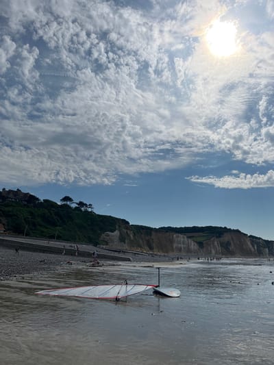 The image depicts a vibrant, sunlit beach scene at low tide, likely during the daytime. The sky is a brilliant blue, dotted with numerous fluffy white clouds, with the sun shining brightly in the upper left corner, casting a warm glow. The beach itself is a mix of pebbles and wet sand, with shallow water reflecting the sky.

In the foreground, a windsurfing board and sail are partially submerged in the shallow water, its white sail accented with red trim. Further back along the beach, a scattering of people are visible, some walking along the shoreline, others gathered in small groups. The beach is backed by a steep, vegetated cliff face, with trees clinging to its slopes. At the base of the cliff, a paved walkway runs parallel to the beach, and a few buildings can be seen nestled among the greenery.

The overall impression is one of a pleasant, active beach day. The bright sun and scattered clouds suggest fair weather, and the presence of people engaged in leisure activities points to a relaxed atmosphere. The cliffs and the architecture hint at a coastal town or village. There is no visible text in the image.