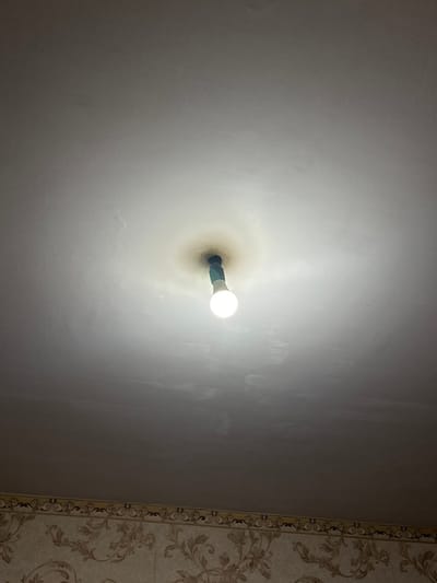 The image captures an upward-facing view of a single, illuminated light bulb mounted directly into a light-colored ceiling or wall. The light bulb is a modern, energy-efficient type with a distinct white, rounded globe and a dark-colored (possibly green or blue) base where it connects to the fixture. Around the base of the light fixture, there is a prominent, circular area of yellowish-brown discoloration on the ceiling/wall surface, suggesting accumulated dust, heat marks, or age-related staining.
The main surface of the ceiling or wall is a uniform, light color, appearing as off-white or light grey, with subtle textural variations. To the far right of the frame, a vertical strip of patterned wallpaper is visible. This wallpaper features an intricate floral or damask design in muted tones, indicating that the light source is located within a room that has decorative wall coverings. The strong glow from the bulb suggests it is the primary light source in the immediate vicinity. The overall setting is an indoor space, likely a room, illuminated by this single fixture. No other objects, people, or activities are depicted, and no text is visible.