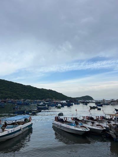 The image shows a harbor filled with numerous boats of various sizes, anchored in calm water. A lush, green, tree-covered hillside forms the backdrop, with rocky outcroppings visible along the shoreline. The sky above is overcast with scattered clouds, some of which are illuminated by sunlight, creating a dappled effect.
In the foreground, several white speedboats are docked together. One of these speedboats has the registration "24K KH-1413" and the words "MINH SON TOUR" visible on its side. Another boat to its right has "PHƯỚC THỊ" written on it. These speedboats appear to be set up for passenger transport, with rows of seating under canopies and life preservers visible.
Further out in the harbor, a larger fleet of traditional fishing boats, mostly blue and white, are moored. These boats vary in size and some have flags attached. In the distance, other islands or landmasses can be seen. The water reflects the sky and the boats, creating a serene, almost still surface. The scene suggests a bustling marine activity area, likely a port or tourist hub. The location is identified as Vinh Hai, Vietnam.