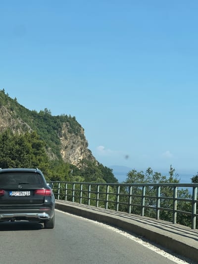 The image depicts a rear view of a black Mercedes-Benz SUV driving on a paved road. The vehicle's license plate reads "KO BN419". The road curves to the right, with a concrete barrier and a metal railing along its edge. Beyond the railing, a steep, rocky cliff covered in dense green vegetation rises on the left side of the frame. The sky is clear and blue. In the distance, a body of water, likely the sea, is visible. The scene appears to be a scenic route, possibly in a coastal or mountainous region. There are no people clearly visible in the image, though a small figure in blue is standing on the vegetation-covered cliff face. The image is taken on a sunny day.
