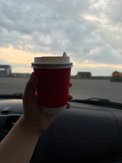 A hand holds a red paper coffee cup with a white lid inside a vehicle. The cup is positioned in front of the windshield, which shows a cloudy sky with the sunset visible through the clouds. In the distance, through the windshield, there appears to be an open area with some buildings and vehicles. The dashboard of the car is visible in the foreground. The setting appears to be outdoors, possibly in an open area near a road or parking lot, given the presence of buildings and vehicles in the background. The overall lighting suggests it is either sunrise or sunset. No specific landmarks or identifying features of Ust-Syny, Russia are discernible in the image.