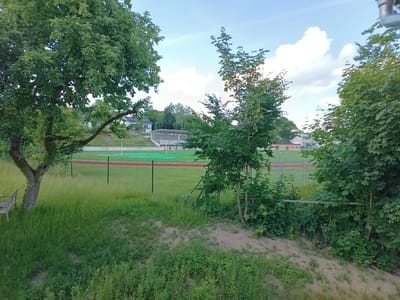 The image shows an outdoor view of a sports field in Oberviechtach, Germany. A red running track surrounds a green field, likely a soccer field. A fence separates the field from a grassy area in the foreground, where a tree and a park bench are visible. In the background, a building with a covered seating area stands behind the field, and residential buildings are situated on a hill. The sky is blue with scattered white clouds.