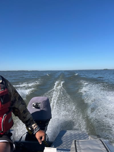 A man wearing a camouflage long-sleeved shirt, a red life vest, and black shorts, is steering a boat on a body of water under a clear blue sky. He is holding the steering mechanism of the boat's outboard motor with his right hand. The motor is covered with a gray canvas. Water is churning and splashing behind the boat, creating a wake. In the distance, a thin line of trees is visible on the horizon, suggesting land. The image appears to be taken from the perspective of someone sitting behind the person steering. The location is not specified in the image or provided information beyond it being in the city of Beregovoe, Russia.