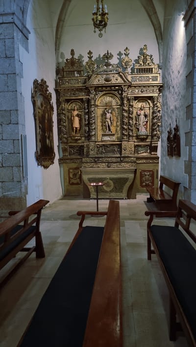 This image shows the interior of a building, likely a church or chapel, in Cadaqués, Spain. The main subject is an ornate, gilded altarpiece with detailed carvings and religious figures. Below the altarpiece, a framed painting hangs on the wall. Wooden benches with dark upholstered seats are arranged on the stone floor, suggesting a place of worship or quiet contemplation. The lighting appears to be artificial, with a chandelier visible on the left and a candle-like light fixture attached to the altarpiece. The walls are a light color, and the ceiling is arched, typical of older architectural styles. The stone floor is made of large, rectangular tiles. The overall impression is one of historical and religious significance, with a somber and peaceful atmosphere. There are no people visible in the scene, nor any indication of the time of day or weather. No text is visible in the image.