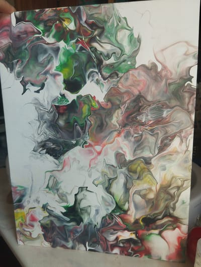 The image displays a close-up, angled view of an abstract acrylic pour painting on a white canvas. The painting features a swirling, marbled pattern created by the flow of various colors, including white, black, gray, various shades of green, pink, yellow, and hints of red. The paint has created organic, fluid shapes with defined edges and some areas with subtle textural effects. The canvas is oriented vertically. The background is out of focus, but appears to be an indoor setting with some soft lighting. There is no discernible connection to Lobnya, Russia within the image itself.