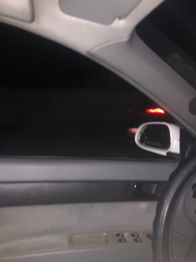 The image shows the interior of a car at night, looking out the driver's side window. The dark exterior is illuminated by the red taillights of another vehicle. A white side-view mirror is visible, reflecting the red lights and the interior of the car. The steering wheel is partially visible on the right side of the frame. The door panel and window controls are also visible. There are no people visible in the image. The scene appears to be on a road within the city of Khujakent, Uzbekistan.