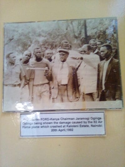 A sepia-toned or black and white photograph, approximately 8x10 inches, is affixed to a plain, light-colored wall. The photograph depicts a group of approximately ten to twelve African adult males in an outdoor setting with dense foliage in the background. In the foreground-center, the interim FORD-Kenya Chairman Jaramogi Oginga Odinga, wearing a light-colored suit, tie, and cap, observes a long, slender, cylindrical object being held horizontally by another man in a light-colored t-shirt and shorts. Other men are clustered around, observing the central figures. The event depicted occurred at Kaloleni Estate, Nairobi, Kenya, on April 20, 1992, as indicated by a white rectangular caption placed directly below the photograph. The physical photograph is located in the city of South Sakwa ward, Kenya.