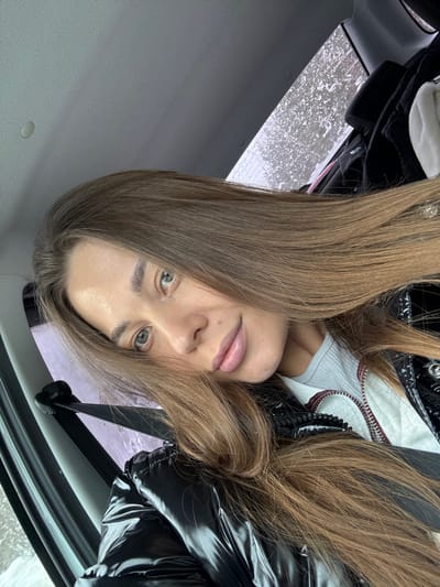 The image captures a female individual taking a selfie inside a vehicle in Bol'shoi Talmaz, Russia. The subject, a young woman with long, light brown hair and green eyes, is in the foreground, looking towards the camera with a slight smile. She wears a light grey ribbed top and a black shiny puffer jacket. A seatbelt is visible across her left shoulder and chest. The vehicle's light gray interior ceiling is visible in the upper frame. Through a rear window in the background, a blurred exterior scene displays white, granular textures consistent with snow or ice, indicative of a cold environment. A folded white item, likely clothing or a blanket, is positioned on a rear surface within the vehicle.