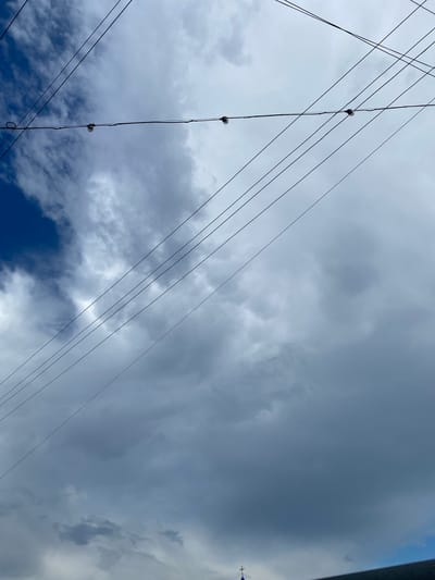 A low-angle outdoor shot shows a dramatic sky filled with a mix of dark gray storm clouds and patches of bright blue sky. Multiple thin, dark electrical wires crisscross the frame, some with small, round light bulbs attached. In the bottom right corner, a dark, sloped roofline is visible, with a faint blue structure topped with a cross peeking out above it. The scene suggests a view from the city of Okhinky, Ukraine, looking upwards towards the sky.