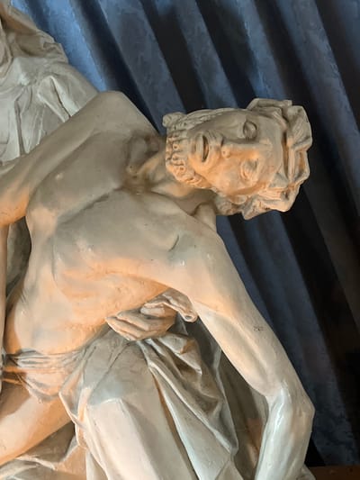 The image shows a close-up of a sculpture of a man, likely depicting a religious figure due to the style and material. The sculpture is made of a light-colored material, possibly plaster or stone, and has a textured surface. The man is depicted with his head tilted back, his eyes closed, and his mouth slightly open, suggesting suffering or repose. His musculature is visible, and he is draped in cloth that folds and creases realistically. The background consists of dark blue, vertically pleated fabric. This sculpture is located in the Église Sainte-Catherine in Honfleur, France, a historic church notable for its shipbuilding-inspired architecture and its collection of religious art.
