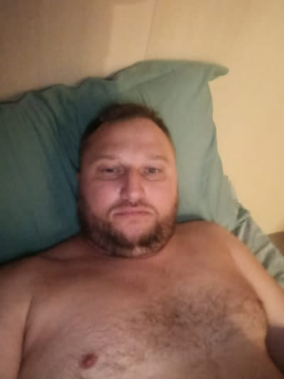 The image shows a close-up, upper body shot of a Caucasian man lying on a light green pillow, with a beige wall behind him. The man has short, dark hair, a beard, and is shirtless. His head is slightly tilted to his left. The lighting is dim and appears to be coming from above. There is no indication of the city of Pisa, Italy, or any other specific location within it.