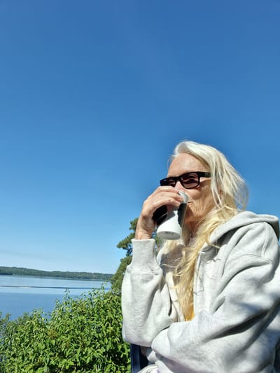 A woman with blonde hair and sunglasses is drinking from a white and black mug. She is wearing a grey hoodie. The background consists of a blue sky, a body of water, and trees. This appears to be a scenic outdoor location.
