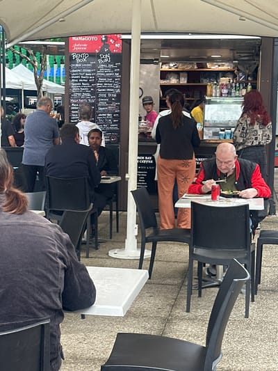 The image depicts an outdoor dining area with several people seated at tables. A food stall with a menu board is visible in the background. In the foreground, a person in a dark jacket is seated at a table, facing away from the camera. To their right, a person in a red and black jacket is eating with chopsticks, a can of Coca-Cola and a plate of food on the table. Further in the background, several other people are visible, some standing and some seated. The environment appears to be a pedestrian area with paved ground and a white umbrella providing shade. The menu board lists Japanese food items, including "Bento Box" and "Donburi". Based on the name "MAMAGOTO Authentic Japanese", and the presence of a Coca-Cola can, this scene could potentially be a food stall in a commercial area of Coffs Harbour, Australia, like the entertainment precinct or a market.
