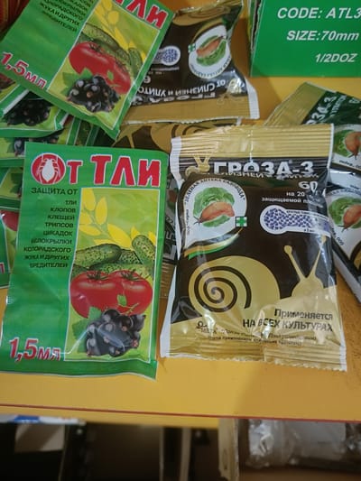 The image shows several packages of garden pest control products arranged on a yellow surface. One prominent package, predominantly green and red, features the text "ОТ ТЛИ" and illustrations of tomatoes, cucumbers, and blackcurrants. Another package, primarily gold and black, displays the text "ГРОЗА-3" with an illustration of a snail. In the background, a green box has text including "CODE: ATL3" and "SIZE: 70mm". The scene appears to be a retail or storage area, possibly a shop or warehouse, with the products laid out for display or organization. There are no people visible in the image. The lighting suggests an indoor environment. The specific location in Berdiansk, Ukraine, cannot be determined from the visual information provided.