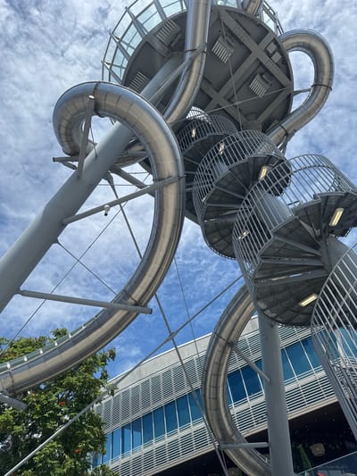 This is a low-angle, close-up shot of a large, metallic amusement park slide structure against a cloudy blue sky. The slide is a winding, tubular structure made of polished metal, supported by a complex network of gray metal beams and columns. Several spiral staircases with metal railings lead up to the platforms of the slide. The overall impression is one of intricate engineering and a playful, adventurous atmosphere. In the background, a modern building with a glass facade and horizontal louvers is visible, partially obscured by the slide structure and a lush green tree. The image appears to be taken in Aventura, Florida, United States of America.