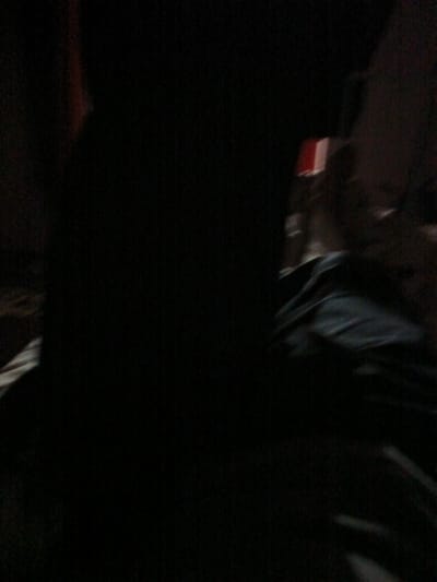 The image is a dark, blurry photograph of a person. The person is mostly obscured by shadow and appears to be wearing dark clothing. There is a hint of red and white in the background, possibly a sign or an object. The overall impression is one of low light and poor visibility, making it difficult to discern specific details about the person or their surroundings. The image does not provide any information about the location in the city of Juma, Uzbekistan.