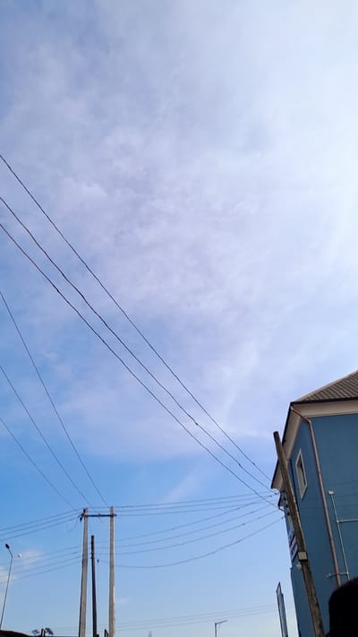 The image shows a view looking upwards toward the sky in Elelenwo, Nigeria. Dominating the upper right corner of the frame is part of a blue building. Numerous power lines stretch across the sky, connecting to utility poles. The sky is a mix of blue and white, partially obscured by clouds.